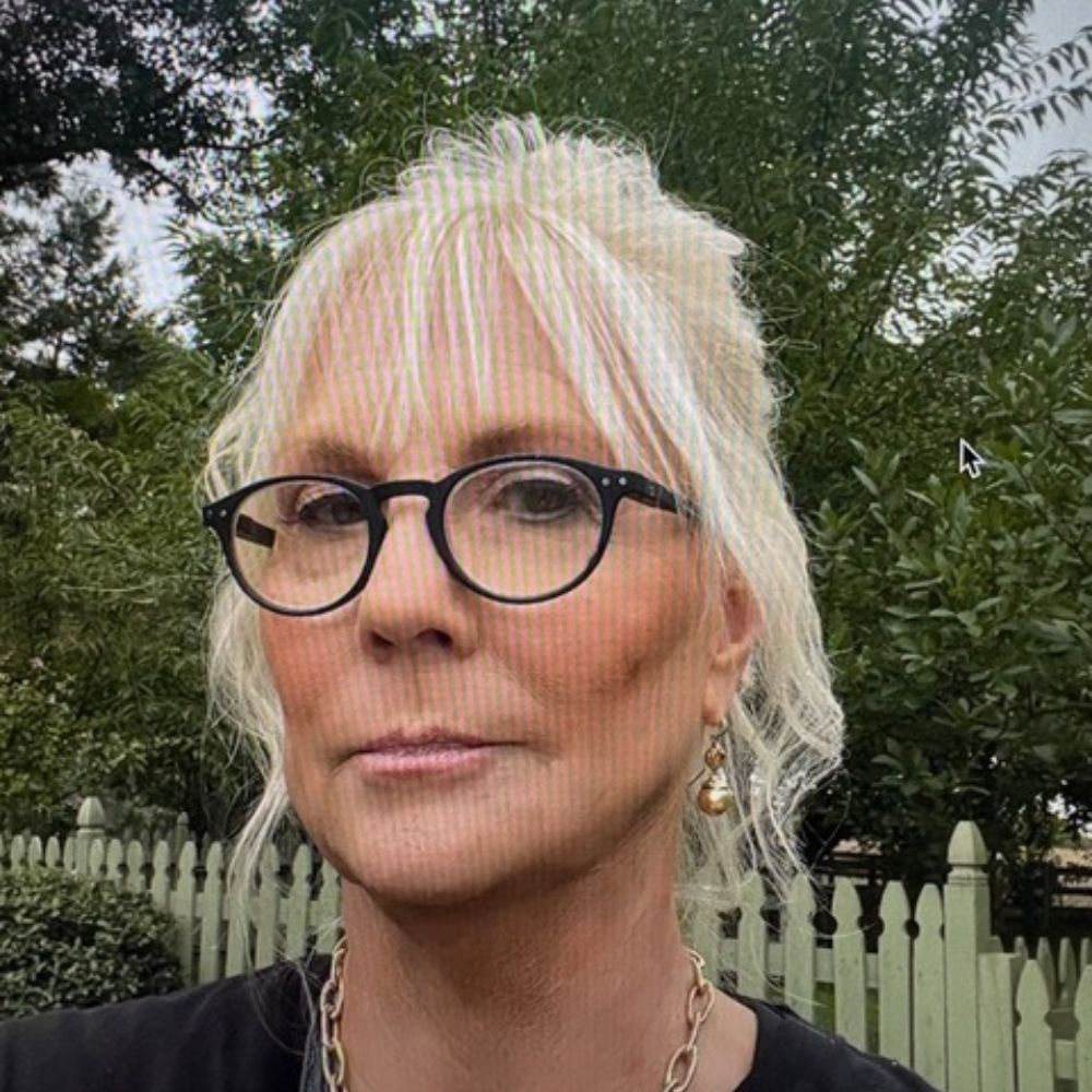 Portrait of a blonde woman wearing round black glasses, gold earrings, and a chunky gold necklace outdoors near a white picket fence with greenery behind her.