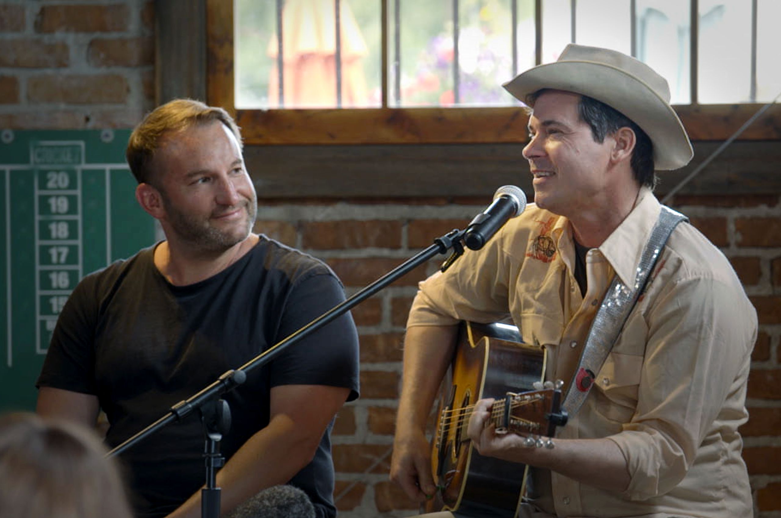 louder-than-guns-2560×1700 - Alexandria Film Festival - Alexandria Film Festival Two men perform live music: one plays guitar and sings into a microphone while the other smiles beside him in a rustic venue with brick walls.